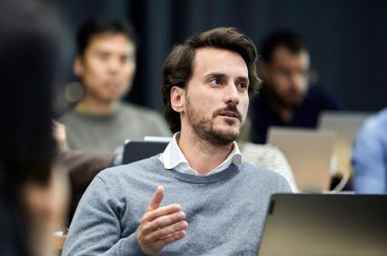 A man in a gray sweater is engaged in a discussion in a classroom setting.