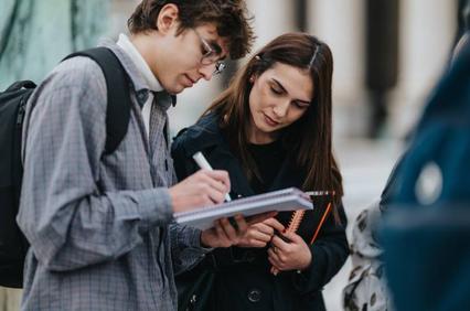 A young man is writing notes while a young woman attentively observes him.