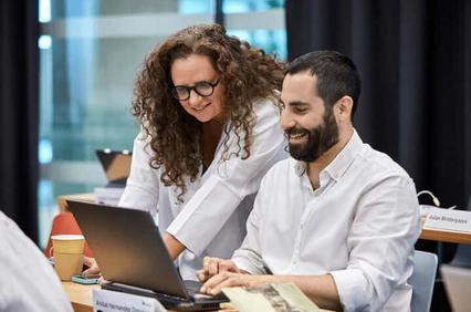 Two professionals, a man and a woman, are smiling and looking at a laptop screen in a modern office setting.