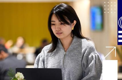 A woman is focused on working on her laptop in a busy office setting.