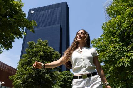 A smiling woman walking among green trees with a modern building in the background.