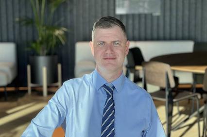 A man in a blue shirt and striped tie smiling at the camera with a modern office background.