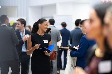 A woman with a name tag smiles and chats at a networking event while holding a notebook.