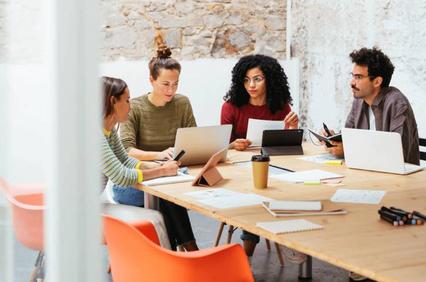 A group of four diverse professionals engaging in a discussion at a cluttered meeting table in a bright workspace.
