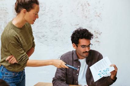 A woman pointing at a document held by a man sitting at a table in a casual office setting.