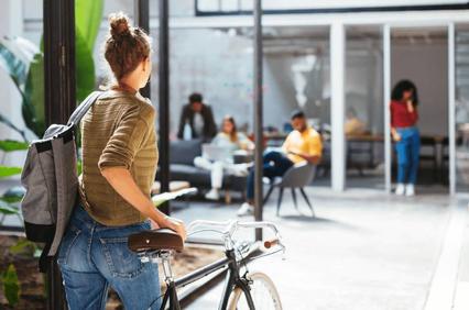 A person with a backpack standing beside a bicycle outside a modern office building, looking at people inside.