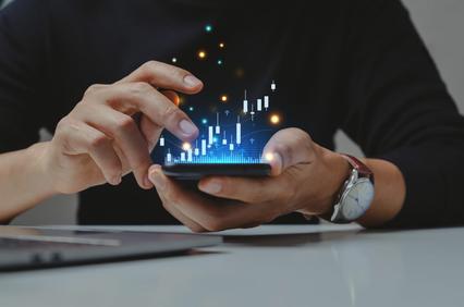 A person analyzing virtual financial data on a smartphone with futuristic holographic statistics, sitting at a desk with a laptop.