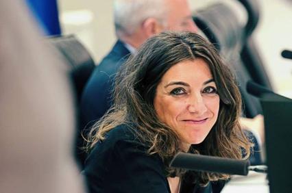 A woman in a professional setting smiles while sitting at a meeting with blurred figures in the background.