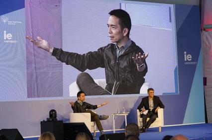 A man gestures while speaking at a conference, with a large screen behind showing his image, as another man listens seated beside him.