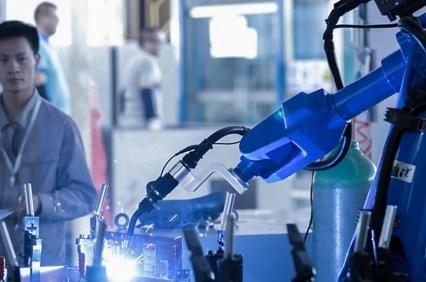 A robotic arm performing a task in a factory setting with a male worker monitoring the process in the background.