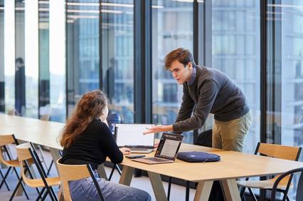 A man is explaining something to a woman at a modern workspace with laptops.
