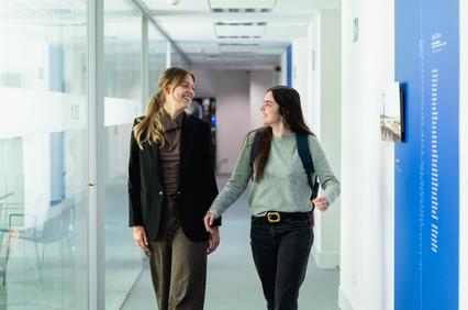 Two women are walking together and smiling in a modern office corridor.