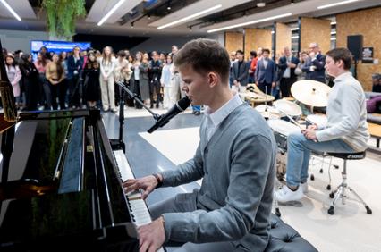 A young man in a grey suit plays the piano at a public event while surrounded by an audience.