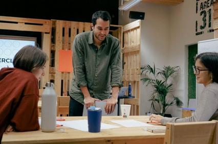 A man stands discussing with two women seated at a table with papers in a casual office setting.