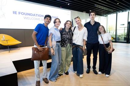 A group of six young adults standing together in a modern lobby in front of a presentation screen that welcomes to the IE Foundation.