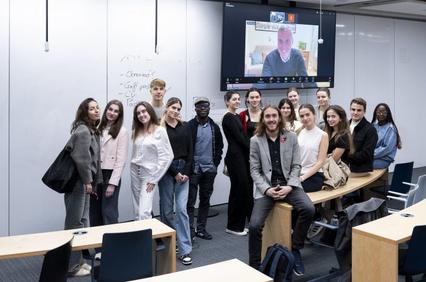 A group of students in a classroom posing for a photo with a teacher appearing on a large screen via video call.