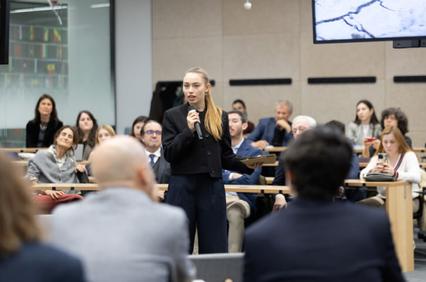 A young woman is speaking into a microphone in front of a group of people seated in a conference room.