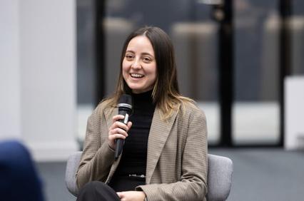 A woman smiles while holding a microphone, sitting and speaking at an event.