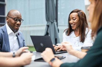 Two professionals, a man and a woman, are smiling and interacting over a laptop at an outdoor table.