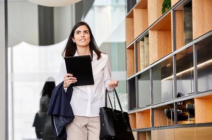 A businesswoman walks through a modern office space carrying a tablet and a handbag.