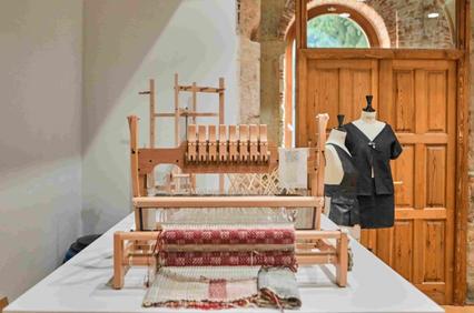 A weaving loom is displayed prominently in a well-lit studio alongside finished garments.