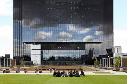 A modern glass building reflecting clouds and people sitting on the grass in front.
