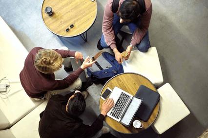 Three people are sitting around a small table in a casual office setting, working together with a laptop and documents.