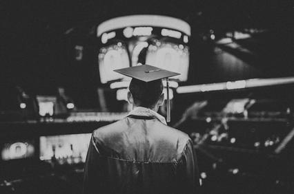 Black and white image of a graduate from behind wearing a cap, facing a blurred stage.