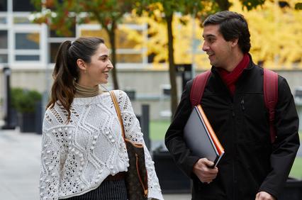 Two people are smiling and chatting while walking on a campus with autumn trees in the background.