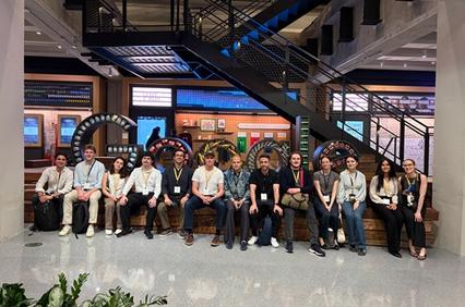 A group of young adults sits together on a bench in a modern interior space with a staircase in the background.