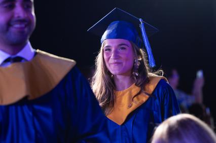 A young woman in a blue graduation gown smiles at a ceremony.