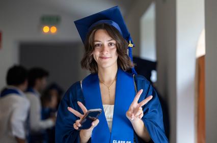 A graduate in a blue cap and gown smiles while making a peace sign and holding a phone.