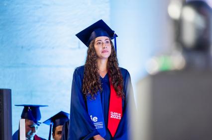 A graduate in a blue gown and cap stands proudly at a graduation ceremony.