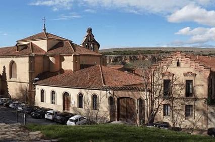 Panoramic view of an ancient monastery complex with terracotta roofs surrounded by sparse vegetation and a clear blue sky.