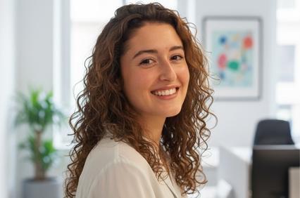 A young woman with curly hair smiles in a bright office environment.