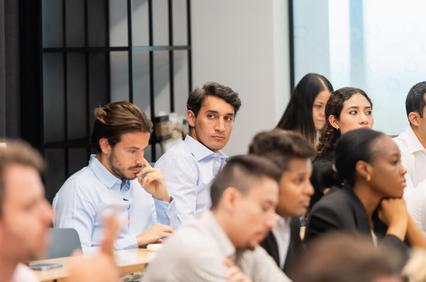 A group of people sitting in a meeting or classroom setting, with a focus on a man in the foreground looking thoughtful.