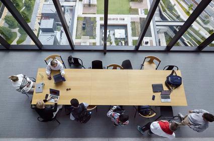 Top view of people sitting around a large wooden table in a modern room with large windows overlooking a green landscape.