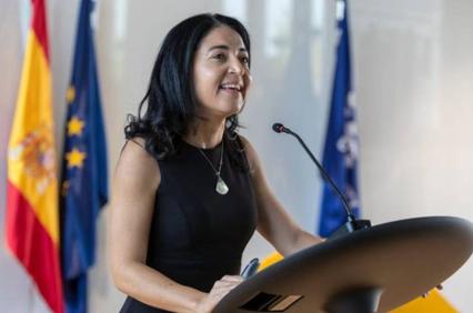 A woman speaks at a podium with various flags in the background.
