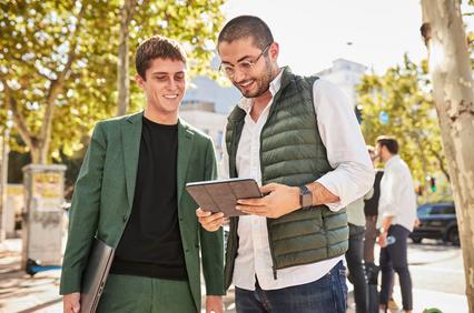 Two young men are discussing something on a tablet outdoors with trees in the background.