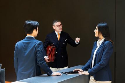 Three business professionals, one man in the center speaking and gesturing to a man and woman on either side, stand in a modern office setting.