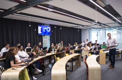 A man presenting in front of an audience in a modern classroom environment.