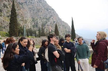 A group of young people listening to a tour guide near a mountainous area.