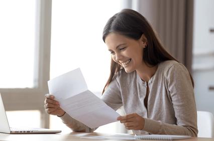 A woman is joyfully reading a letter at a desk with a laptop.