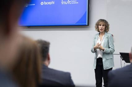 A woman is presenting at a conference with a digital screen behind her displaying logos.