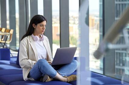 A woman sits comfortably working on her laptop in a modern office setting with large windows.