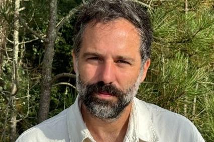 A middle-aged man with a beard and a white shirt standing in front of a wooded area.