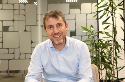 A smiling man in a blue shirt sitting in an office environment with plants and geometric wall patterns.