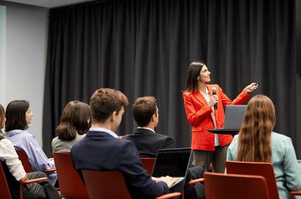 A woman in a red blazer speaks at a podium to an audience in a conference room