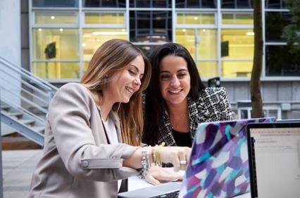 Two women are smiling and working together on a laptop outdoors.