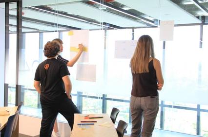 Three people are collaborating as they place papers on a wall in a bright office space.